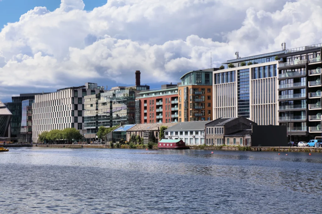 Modern waterfront buildings against a cloudy sky, reflected in a calm urban river.