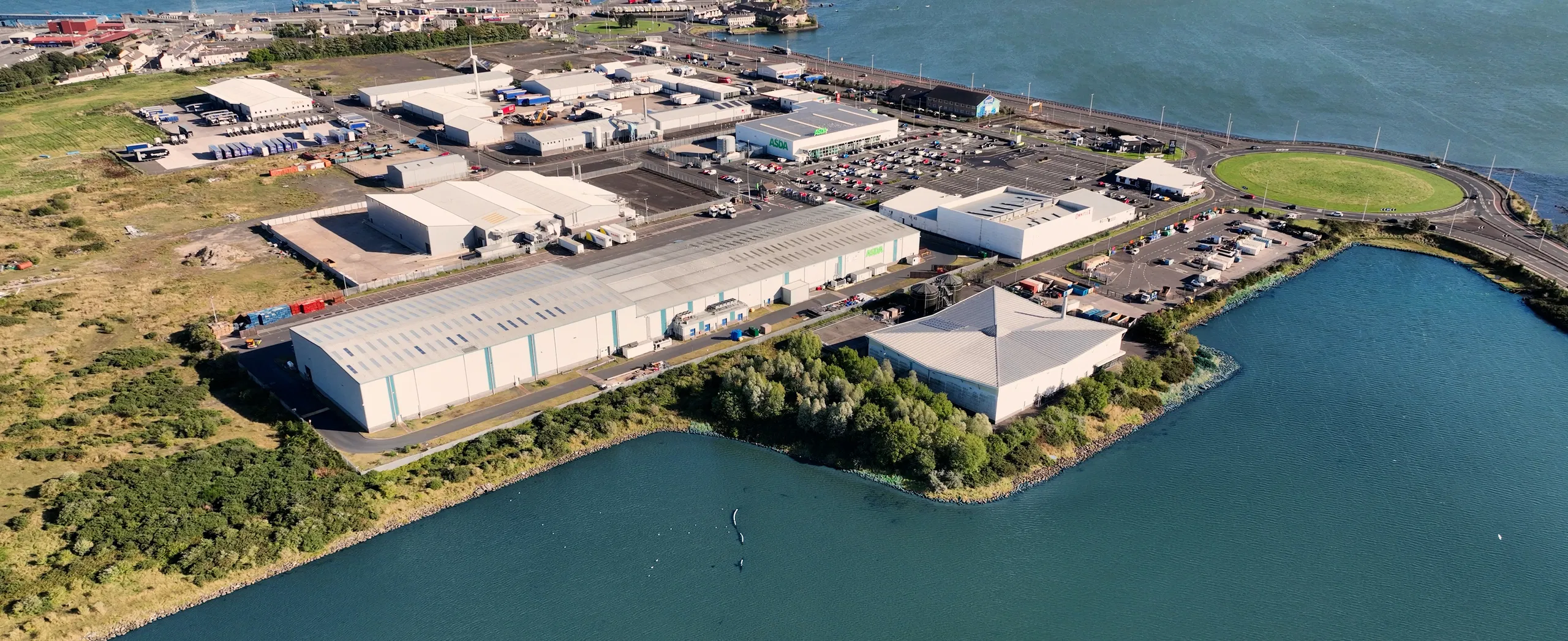Aerial view of industrial warehouses and buildings by a large waterfront, surrounded by greenery and roads.