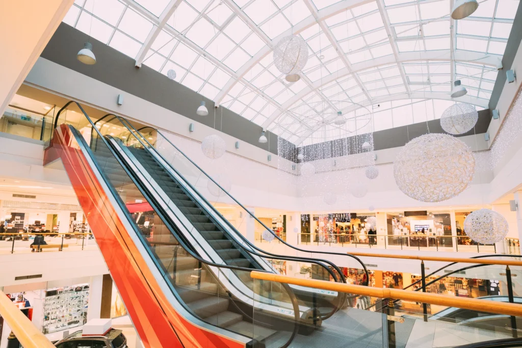 Modern shopping mall interior with escalators and decorative hanging lights under a glass ceiling.