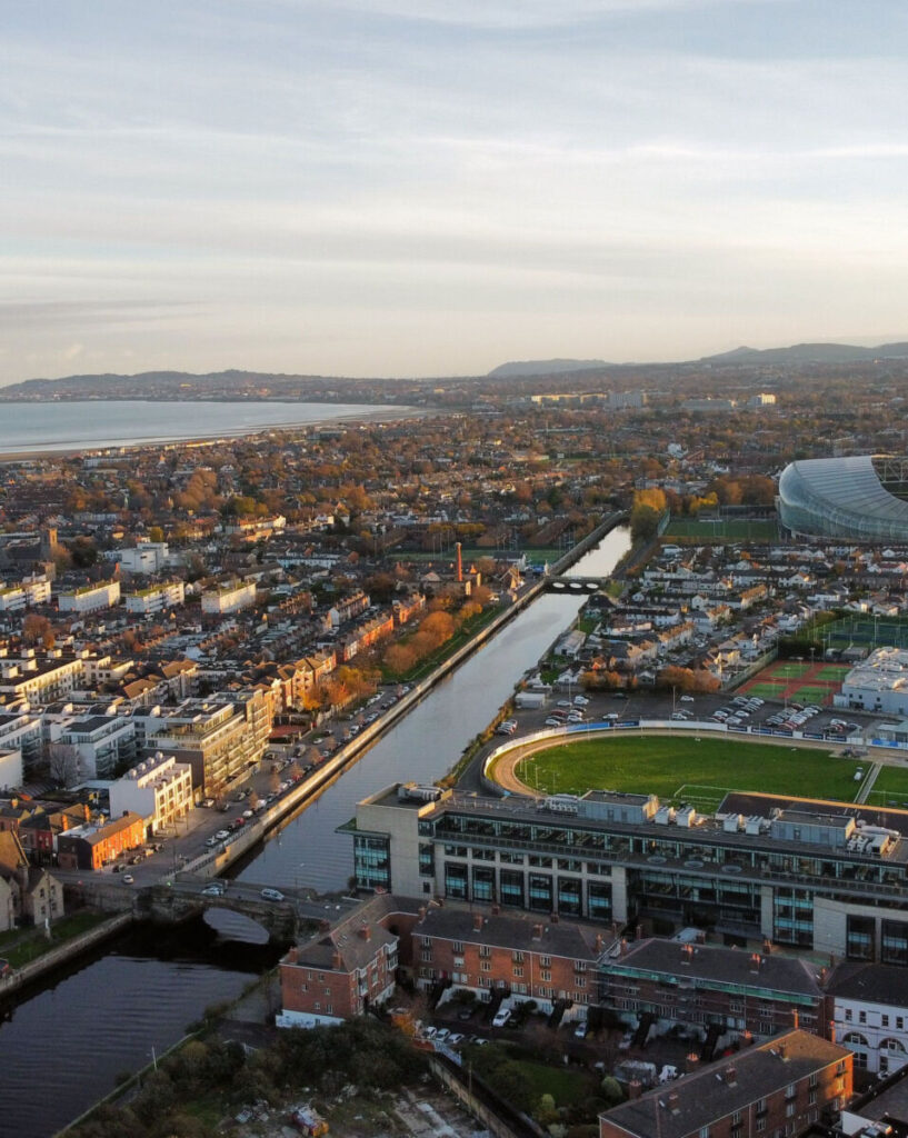 Aerial view of a cityscape with a river, residential areas, and a stadium under a cloudy sky.
