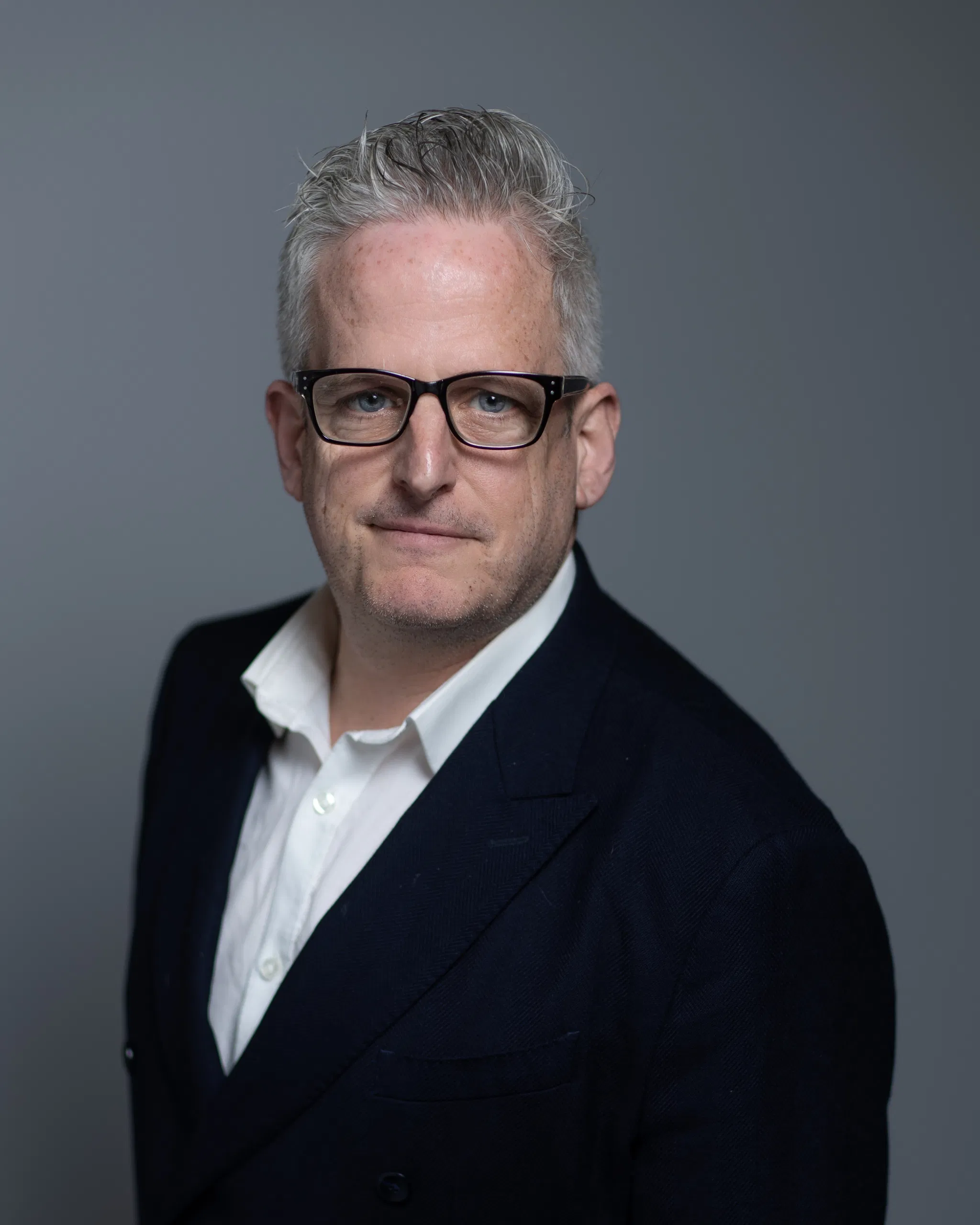 Man in glasses and suit looking confidently at camera, gray background.