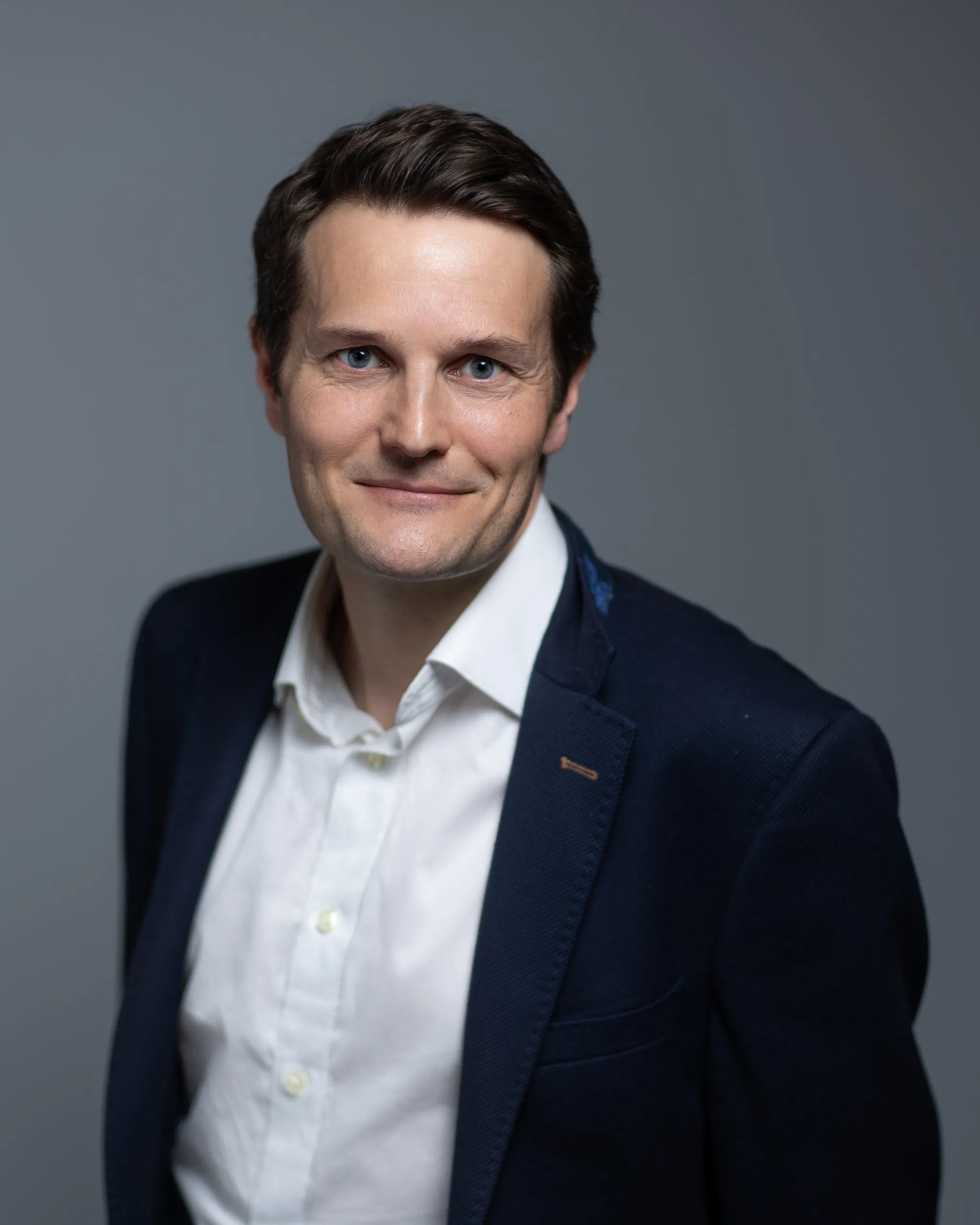 Smiling man in a suit and white shirt against a gray background, looking confidently at the camera.