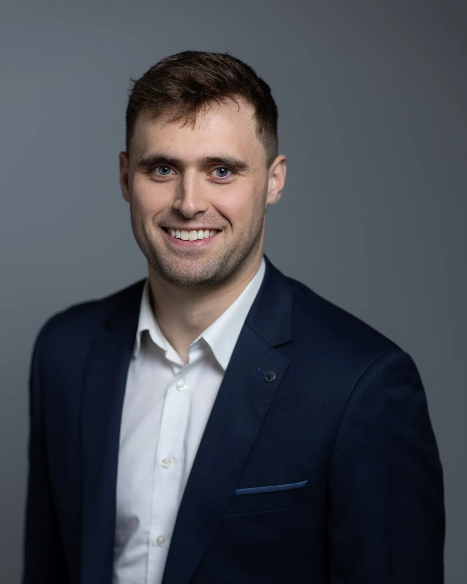 Man in a navy suit and white shirt smiling at the camera against a neutral background.